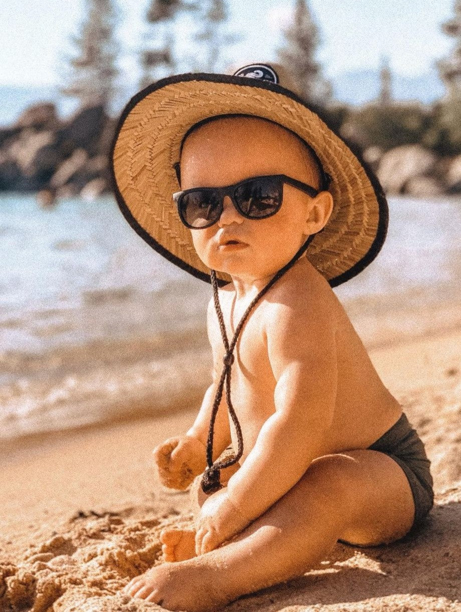 Baby wearing a straw beach sun hat on the beach wearing sunglasses and playing in the sand