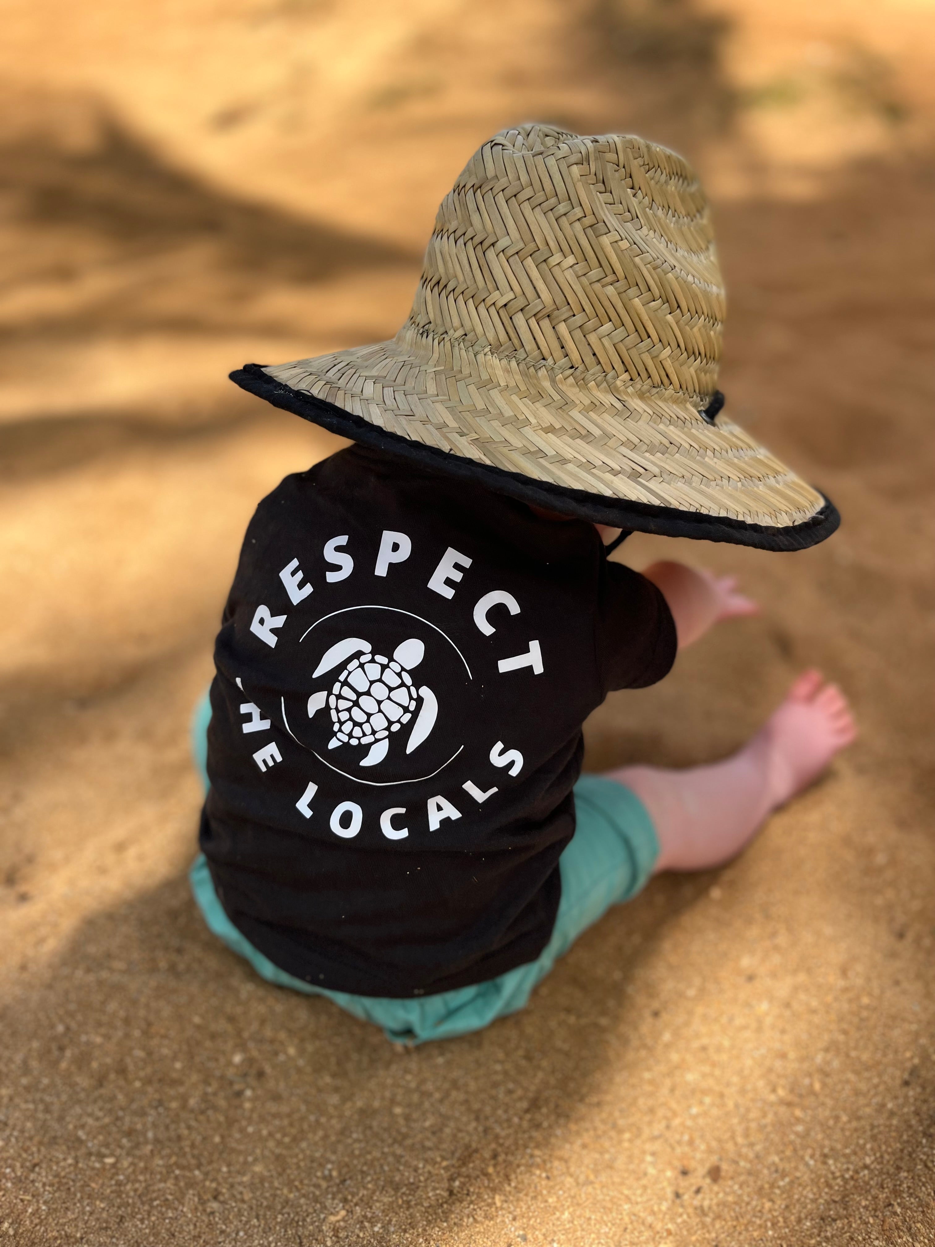 Baby on the beach under some palm trees with a beachy straw sun hat and a stylish respect the local shirt playing in the sand