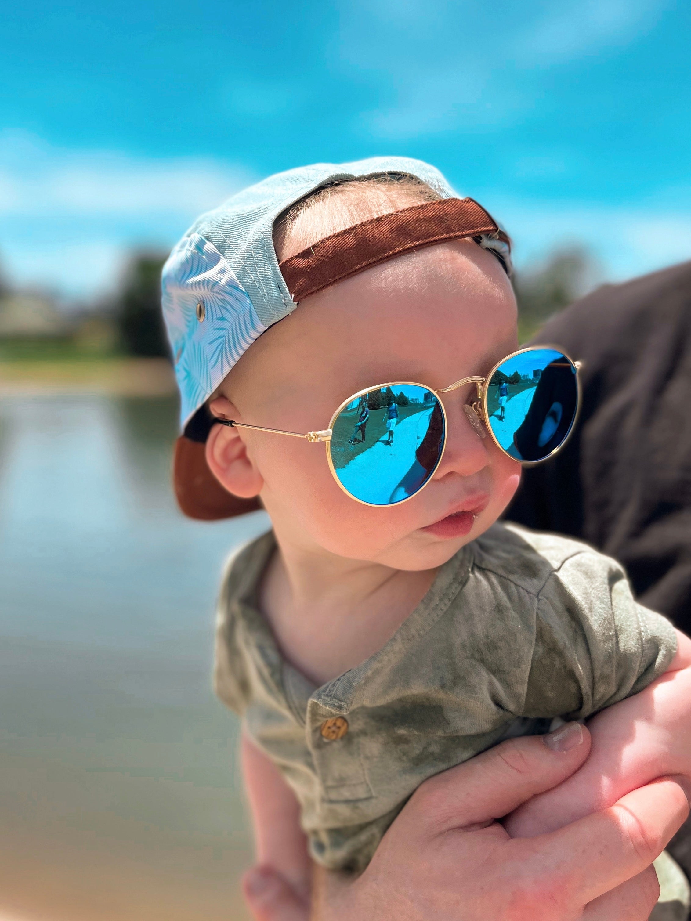 Baby wearing reflective shades in the sunny summer by a lake protecting eyes in a blue snapback 5 panel hat
