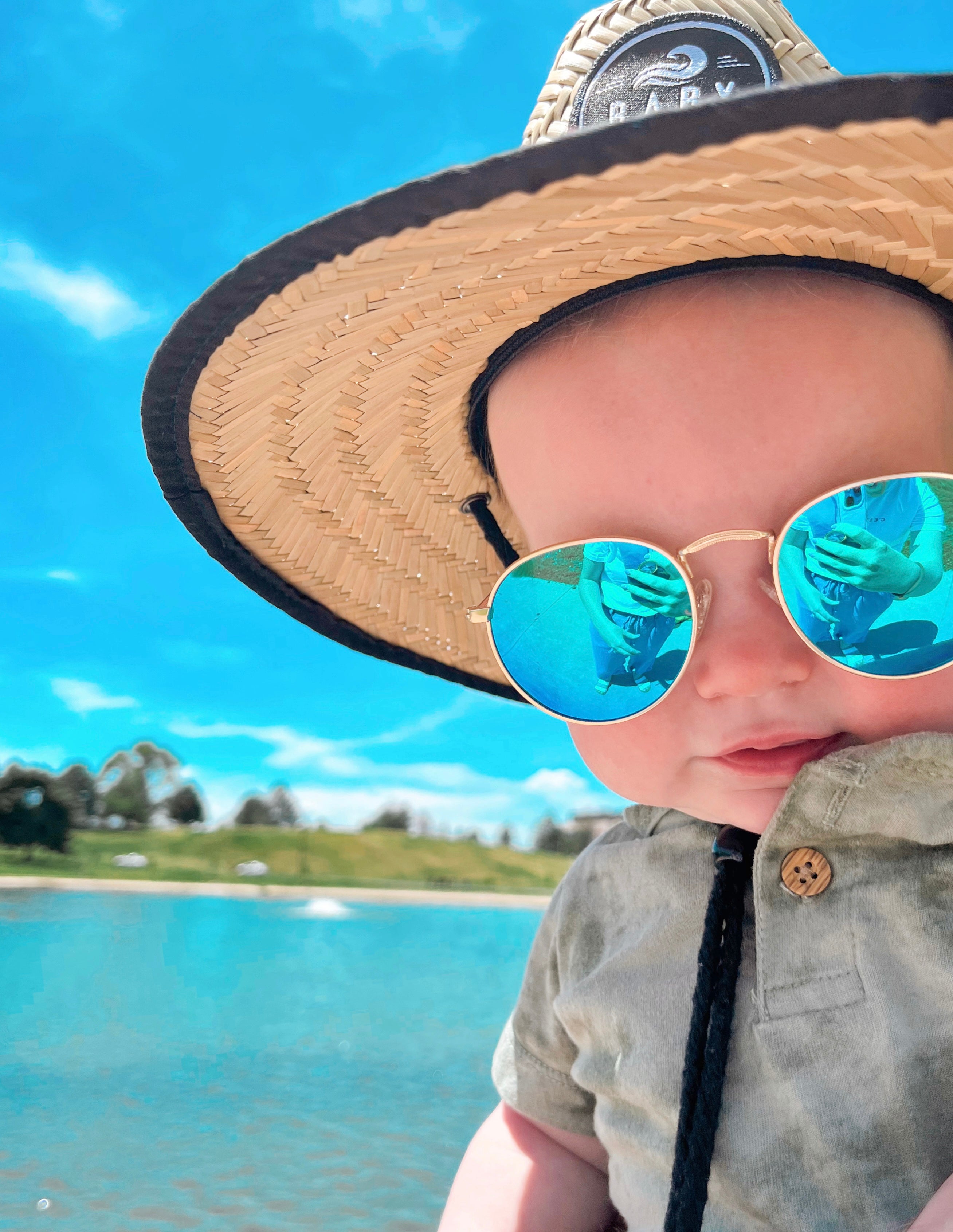 Baby with a sun hat and sunglasses enjoying the sun by a lake with blue reflective lenses