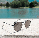 Load image into Gallery viewer, black sunglasses with black rims and lenses to protect babies and toddlers from the sun next to a lake
